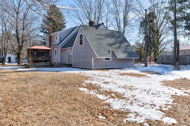 view of snowy exterior featuring a shingled roof, a chimney, fence, and a sunroom