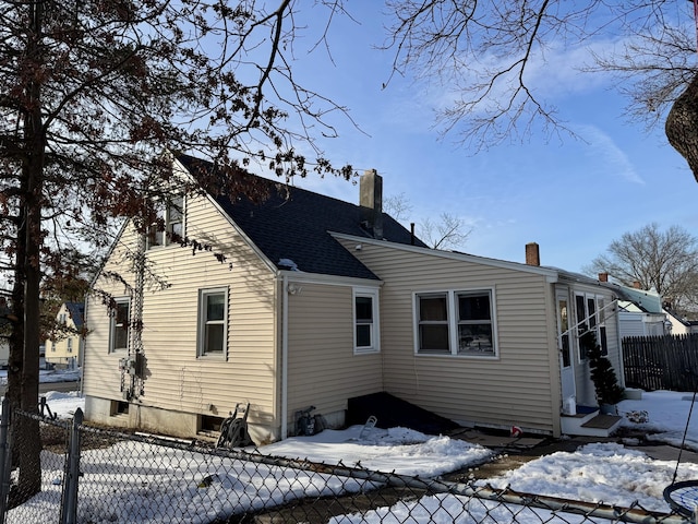 snow covered house with a chimney, fence, and roof with shingles