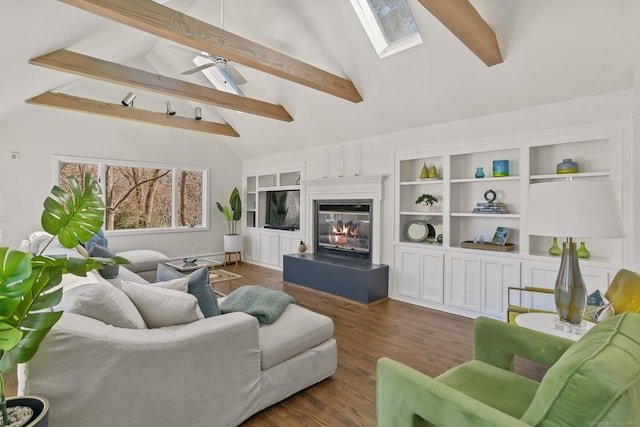 living area with ceiling fan, dark wood-style flooring, a skylight, beam ceiling, and a glass covered fireplace