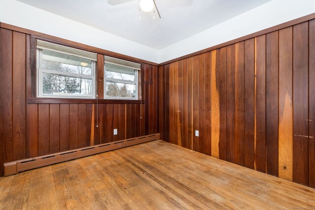 empty room featuring wood-type flooring, ceiling fan, wooden walls, and baseboard heating