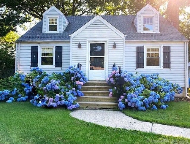 new england style home featuring entry steps, roof with shingles, and a front lawn