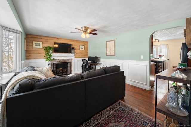 living area with arched walkways, a fireplace, dark wood finished floors, ceiling fan, and wainscoting