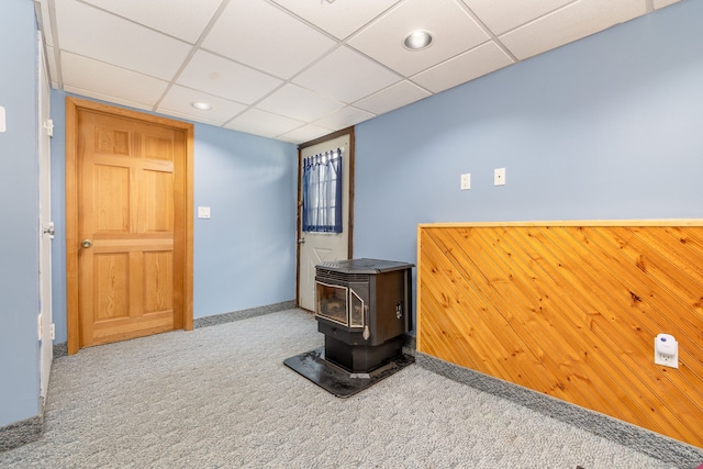 interior space featuring a paneled ceiling, wood walls, carpet flooring, and a wood stove