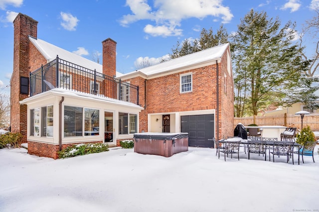 snow covered property featuring brick siding, a hot tub, fence, a balcony, and a garage