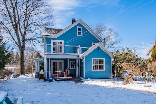 snow covered property featuring a balcony, french doors, covered porch, and a chimney