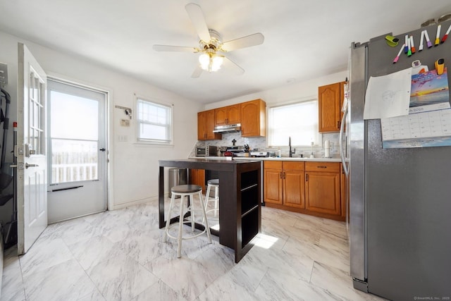 kitchen with a sink, under cabinet range hood, a kitchen breakfast bar, stainless steel appliances, and brown cabinetry