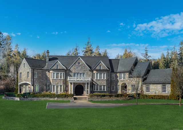 view of front of house with a balcony, stone siding, a chimney, a high end roof, and a front lawn