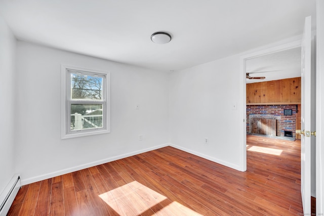 empty room with a brick fireplace, a baseboard heating unit, baseboards, and wood finished floors