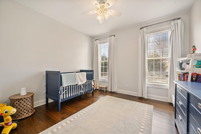 bedroom with a nursery area, dark wood-style flooring, a ceiling fan, and baseboards