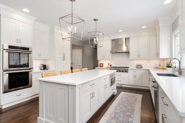 kitchen featuring wall chimney exhaust hood, a kitchen island, appliances with stainless steel finishes, light countertops, and a sink