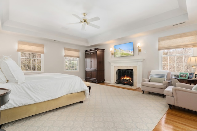 bedroom featuring a raised ceiling, visible vents, light wood-style flooring, a ceiling fan, and a lit fireplace