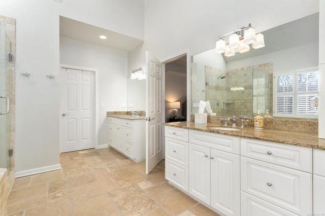 full bath with stone tile floors, two vanities, a sink, baseboards, and a shower stall