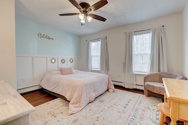 bedroom featuring a ceiling fan, wood finished floors, baseboard heating, and wainscoting