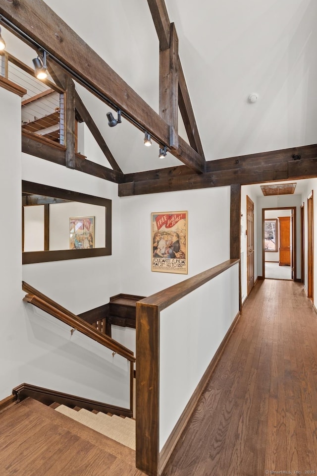 corridor featuring rail lighting, lofted ceiling with beams, baseboards, and hardwood / wood-style flooring