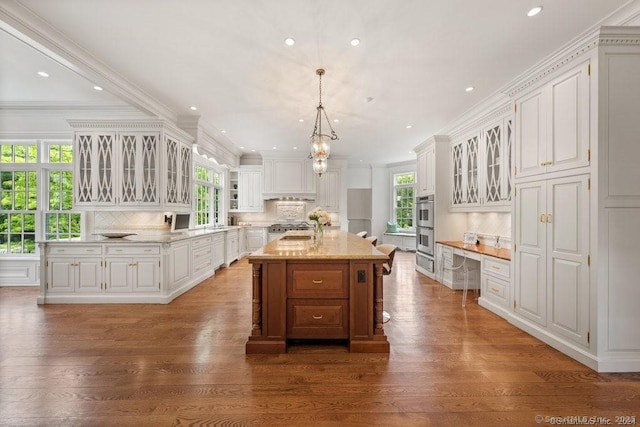 kitchen featuring glass insert cabinets, a kitchen island with sink, white cabinetry, built in desk, and decorative light fixtures