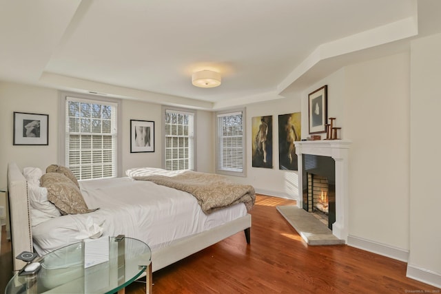 bedroom with a warm lit fireplace, baseboards, a raised ceiling, and wood finished floors