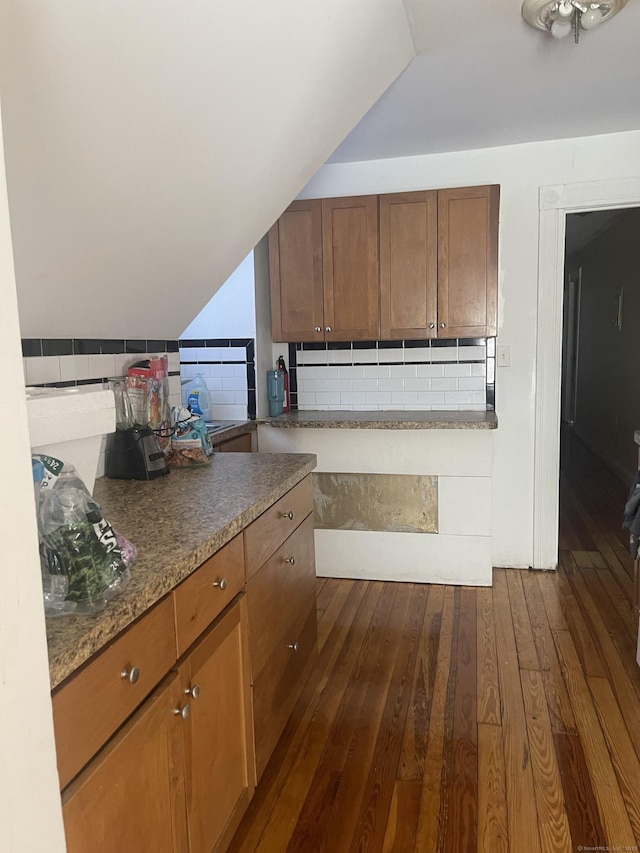 kitchen featuring lofted ceiling, decorative backsplash, dark wood-style floors, brown cabinetry, and dark stone countertops