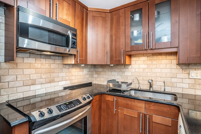 kitchen featuring brown cabinetry, dark countertops, glass insert cabinets, appliances with stainless steel finishes, and a sink