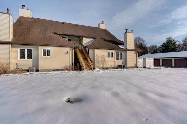 snow covered rear of property featuring a garage, central AC, stairway, and a chimney