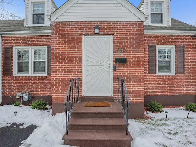 snow covered property entrance featuring an attached garage, a shingled roof, and brick siding