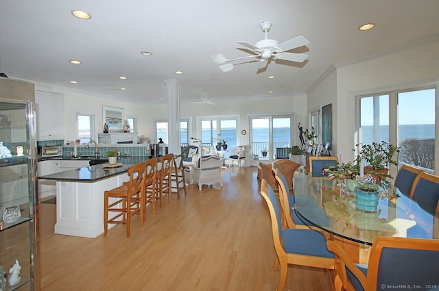 dining room with a water view, a ceiling fan, light wood-style floors, ornamental molding, and decorative columns