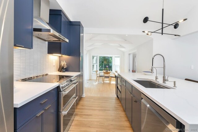 kitchen featuring stainless steel appliances, a sink, wall chimney range hood, blue cabinetry, and light wood-type flooring