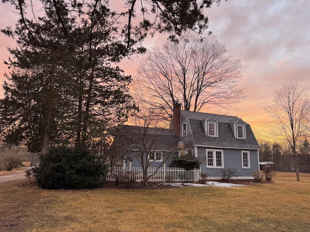 back of house at dusk featuring fence, a gambrel roof, a yard, roof with shingles, and a chimney