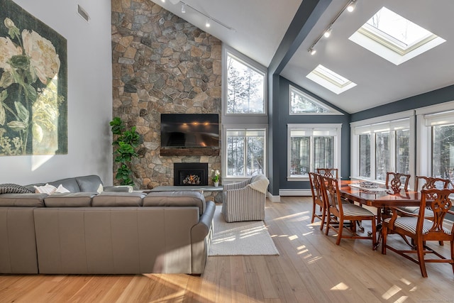 living room with light wood finished floors, a skylight, baseboard heating, a stone fireplace, and high vaulted ceiling