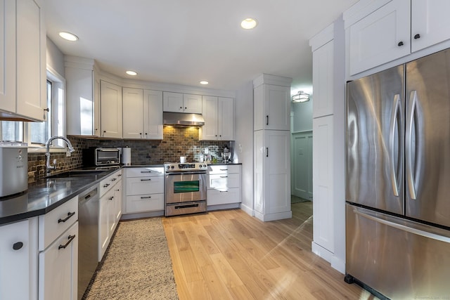 kitchen featuring stainless steel appliances, dark countertops, a sink, light wood-type flooring, and under cabinet range hood