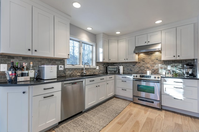 kitchen with dark countertops, light wood-style flooring, stainless steel appliances, under cabinet range hood, and a sink