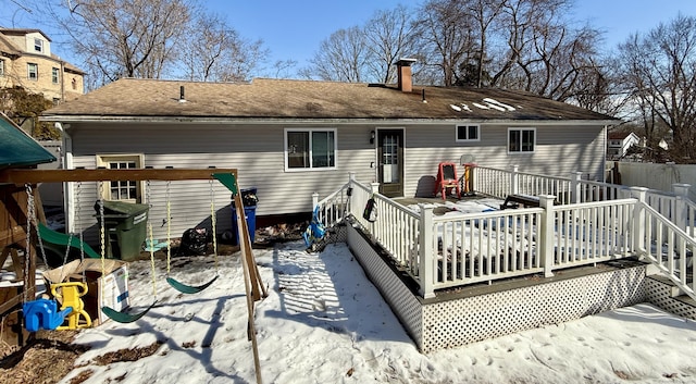 snow covered back of property featuring a deck, a chimney, and a playground