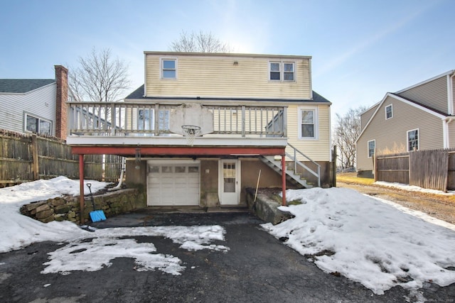 snow covered rear of property with an attached garage, stairs, fence, and aphalt driveway