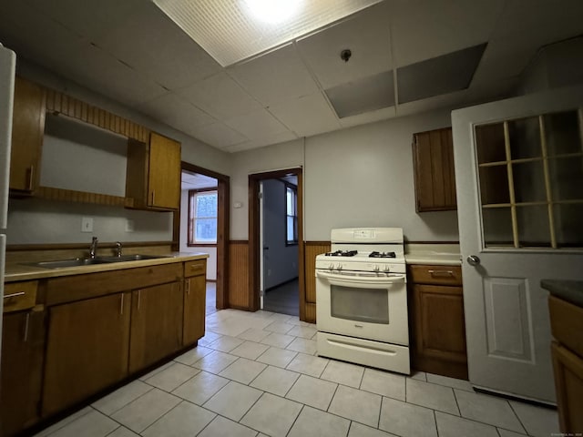 kitchen with light tile patterned floors, white gas stove, a sink, light countertops, and wainscoting