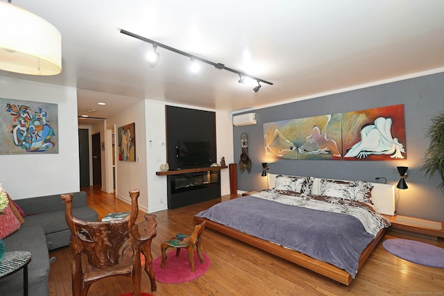 bedroom featuring a wall unit AC, baseboards, wood finished floors, and a glass covered fireplace