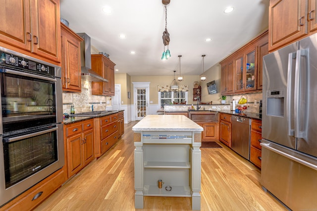 kitchen featuring light wood-style flooring, a peninsula, appliances with stainless steel finishes, brown cabinets, and wall chimney exhaust hood