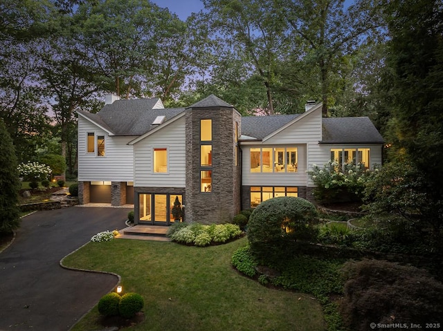 view of front of house featuring a shingled roof, stone siding, a chimney, aphalt driveway, and a front lawn