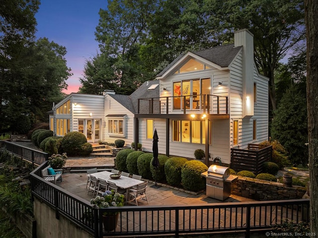 back of house at dusk featuring a patio, a balcony, french doors, roof with shingles, and a chimney