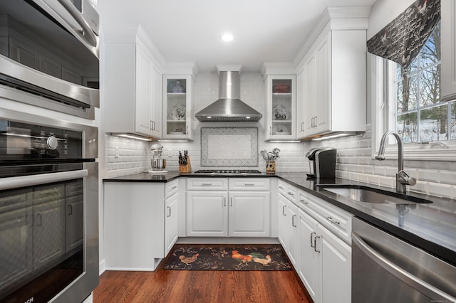 kitchen featuring dark countertops, stainless steel appliances, wall chimney range hood, white cabinetry, and a sink