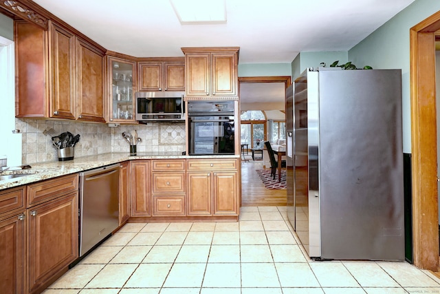 kitchen with light stone counters, tasteful backsplash, brown cabinetry, glass insert cabinets, and black appliances