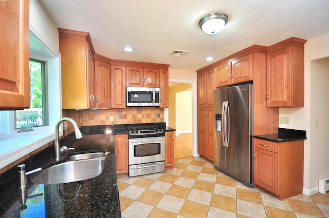 kitchen featuring appliances with stainless steel finishes, dark stone counters, decorative backsplash, a sink, and brown cabinetry