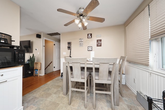 dining space featuring light wood finished floors, ceiling fan, visible vents, and a wainscoted wall