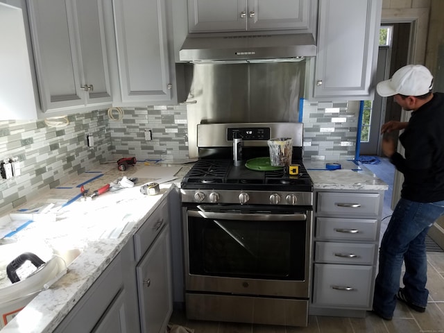 kitchen featuring tasteful backsplash, light stone counters, stainless steel gas range, gray cabinetry, and exhaust hood