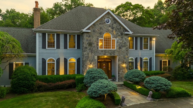 colonial home with a shingled roof, stone siding, a yard, and a chimney