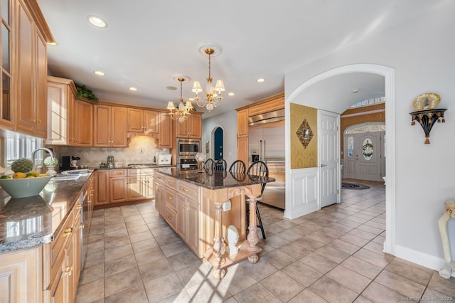 kitchen with built in appliances, arched walkways, a sink, an island with sink, and dark stone countertops