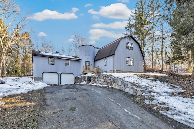 snow covered back of property featuring driveway, an attached garage, and a gambrel roof