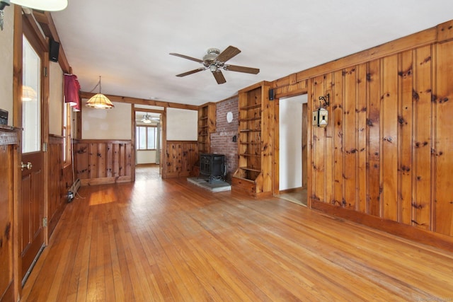 unfurnished living room featuring ceiling fan, wood walls, baseboard heating, light wood finished floors, and a wood stove