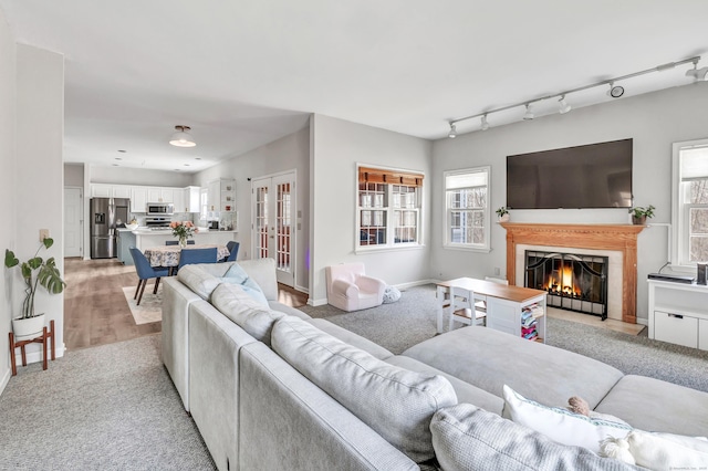 living room featuring a warm lit fireplace, baseboards, light colored carpet, french doors, and track lighting