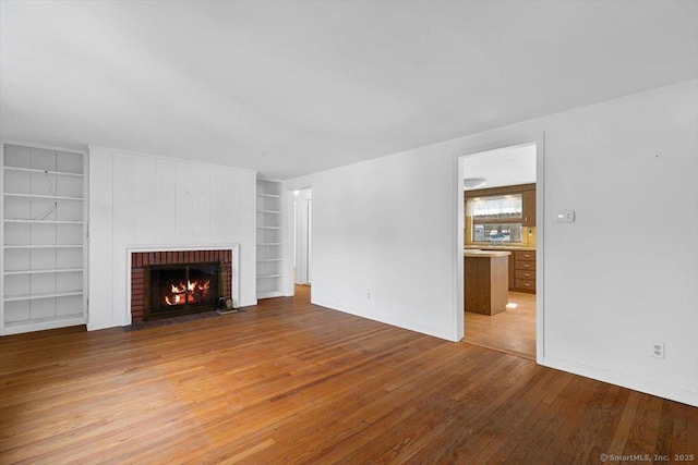 unfurnished living room featuring light wood finished floors, built in shelves, a brick fireplace, and baseboards