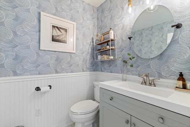 half bath featuring a wainscoted wall, vanity, toilet, and wallpapered walls