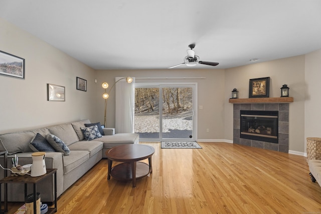 living area featuring ceiling fan, a tiled fireplace, light wood-style flooring, and baseboards